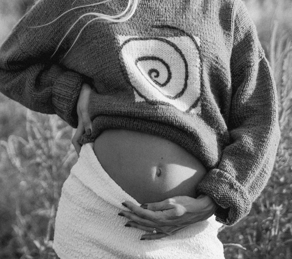 Pregnant mother cradling her bare belly while wearing a knit sweater with a large spiral design.
