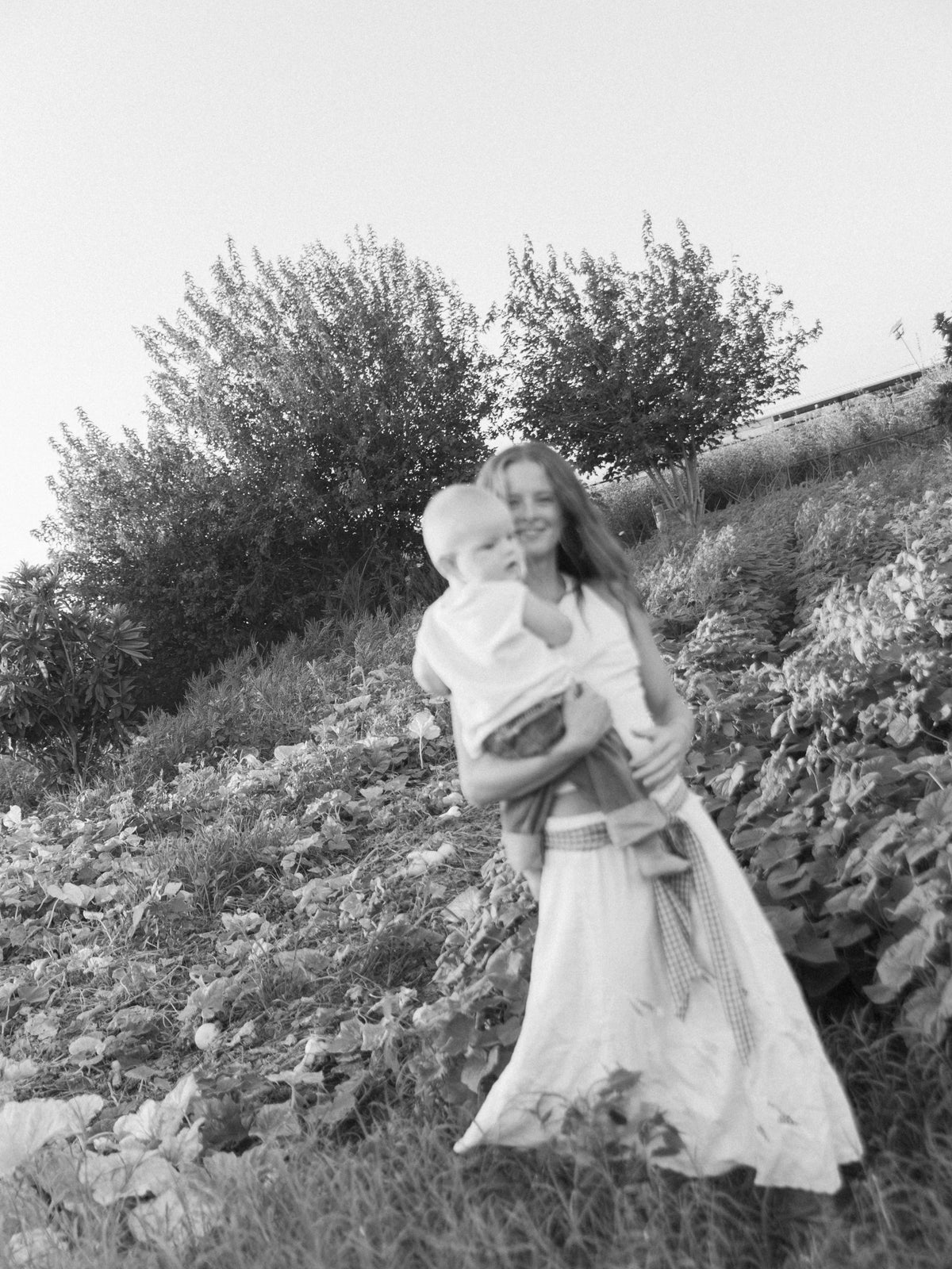 Woman holding a baby while standing among garden plants, photographed in black and white.