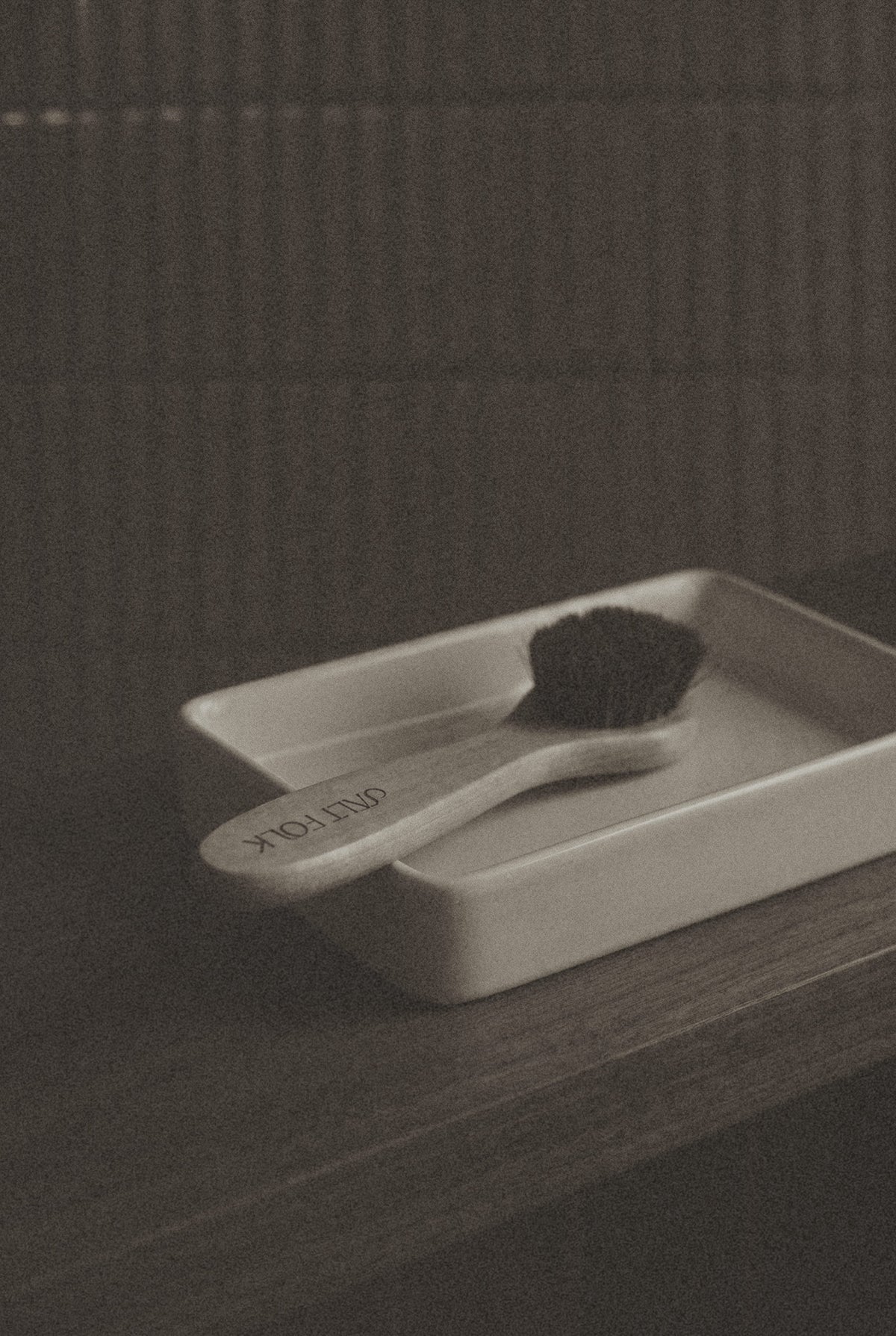 A small white rectangular soap dish holding a toothbrush with a white handle and dark bristles, set on a dark wooden surface against a textured dark background.
