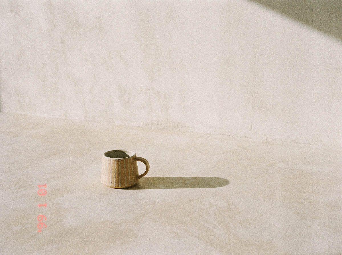 A small beige ceramic mug with a handle, placed on a textured concrete floor against a matching concrete wall, casting a long shadow.