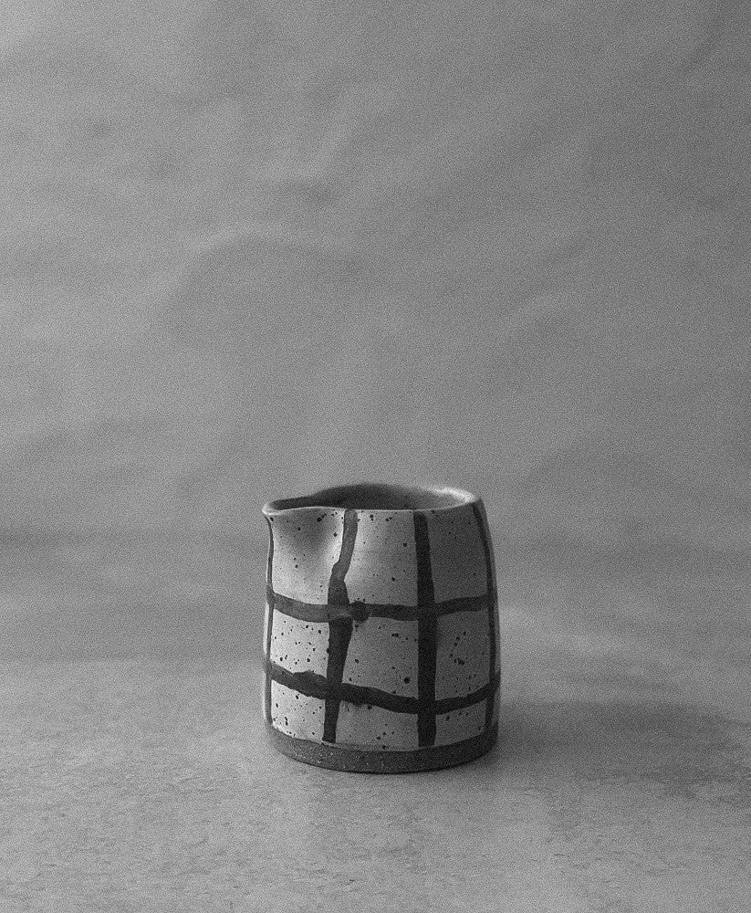 A black and white photo of a small handmade ceramic milk or creamer pitcher with a textured, speckled body and a grid pattern, set against a neutral gray background.