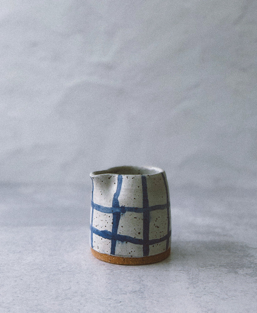 A small handmade ceramic milk or creamer pitcher with a blue grid pattern on a speckled off-white glaze, set against a neutral gray background.