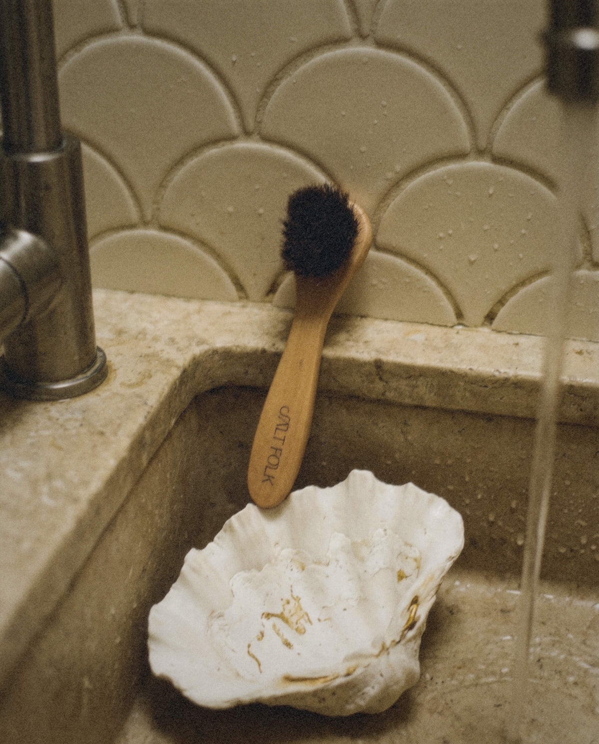 Salt Folk wooden facial dry brush with natural bristles resting in a stone sink beside a white seashell.