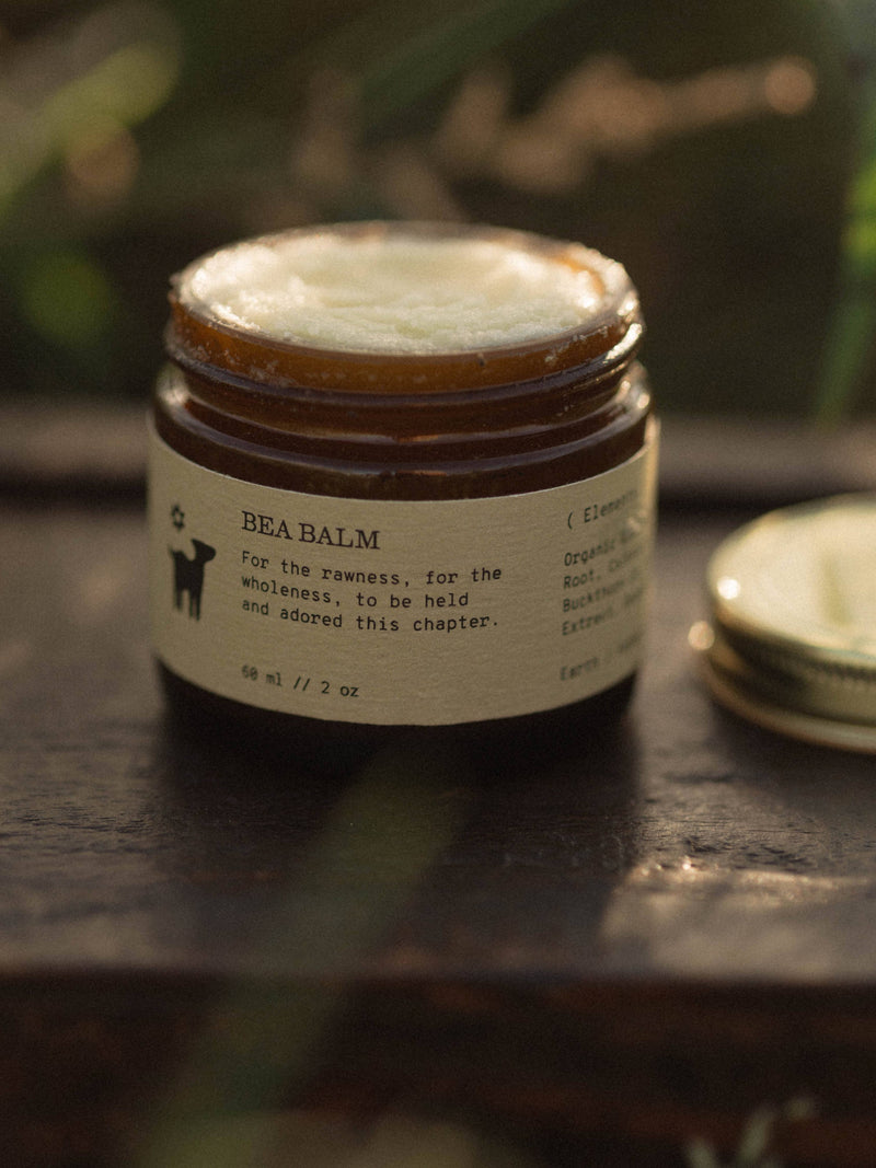 Open amber jar of creamy white Bea Postpartum Balm with its gold lid beside it on a dark wooden surface.