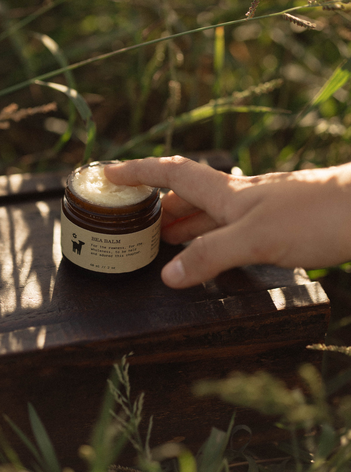 A hand dips a fingertip into the creamy surface of an open amber glass jar of Salt Folk Bea Balm, resting on a wooden surface among sunlit greenery.