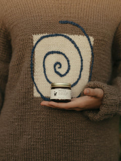 A person wearing a brown knit sweater with a large cream and blue spiral graphic on the chest holds Salt Folk Wren Balm with a gold lid and a white label in their hand.