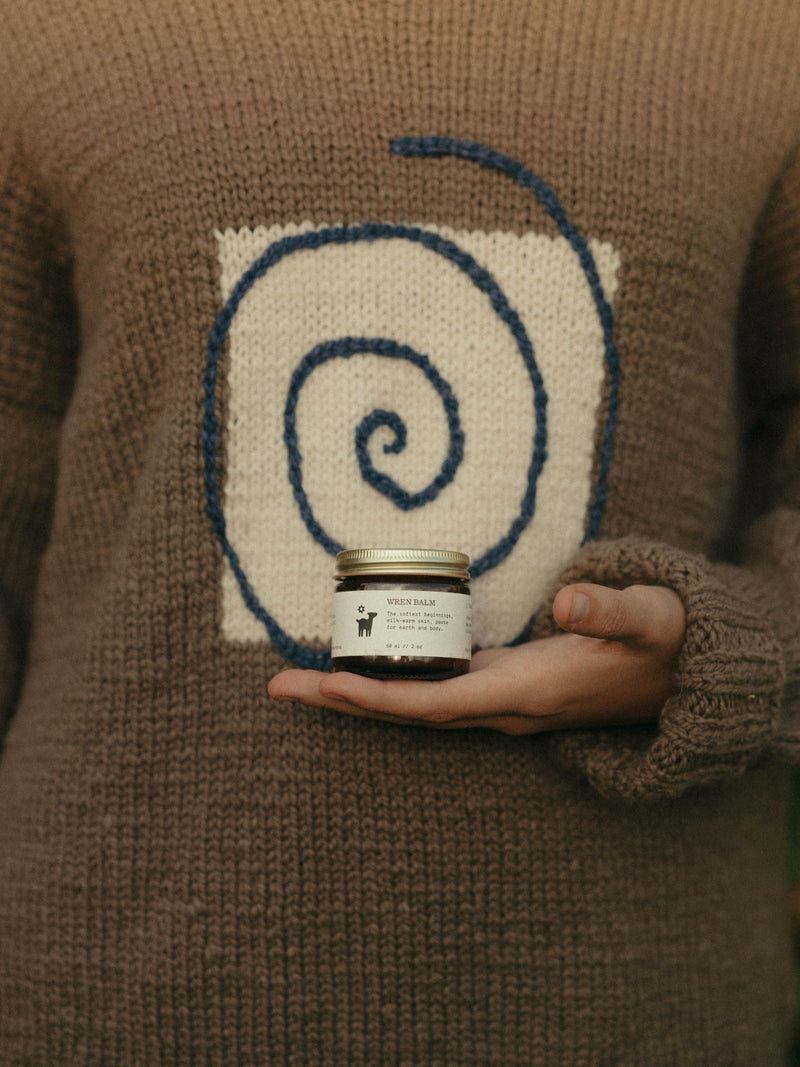 Hand holding a jar of Wren Baby Balm against a brown knit sweater featuring a large blue spiral design.
