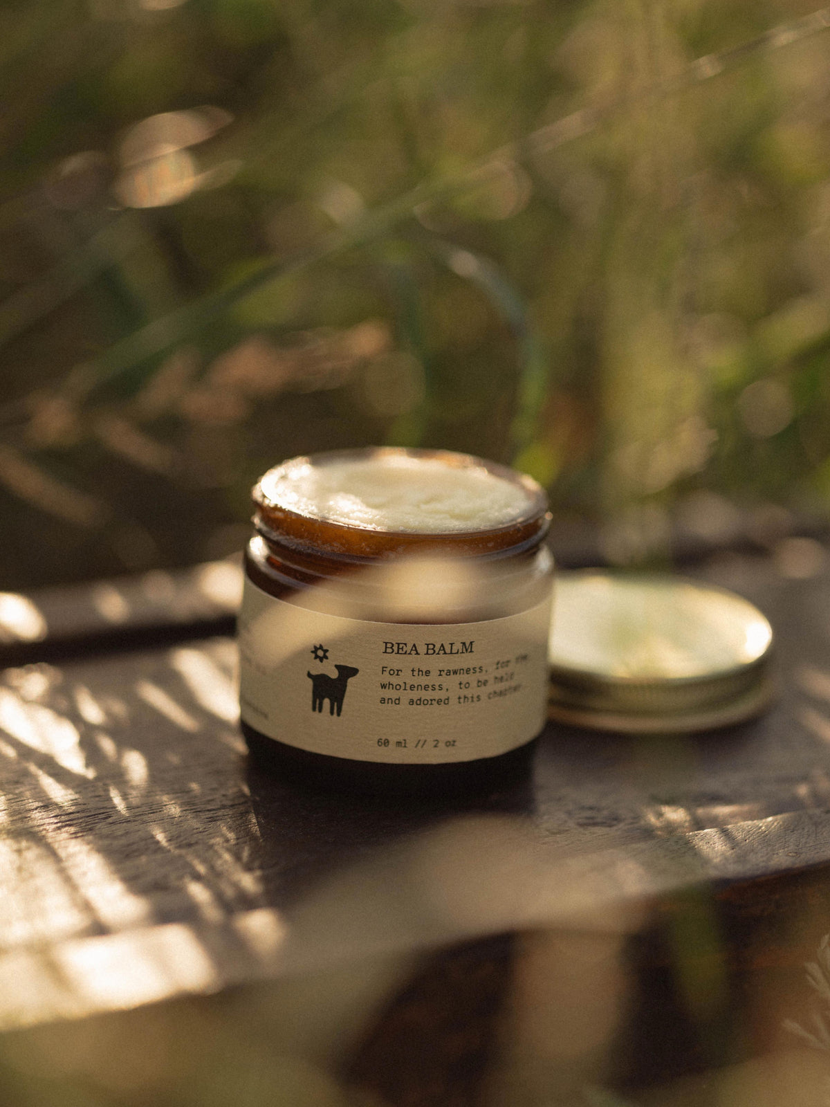 A small glass jar of Salt folk Bea Balm with a metal lid, placed on a wooden surface outdoors with soft natural light and blurred greenery in the background.