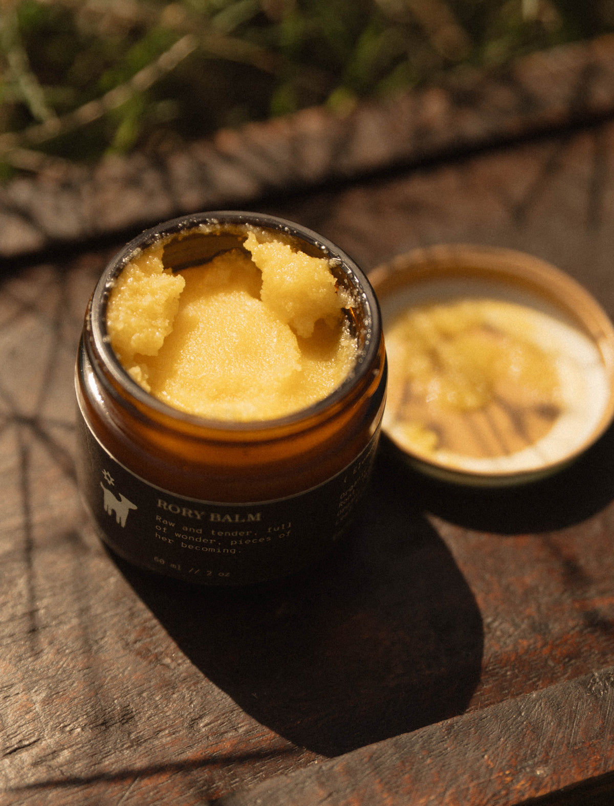 An open amber glass jar of body balm, Salt Folk's Rory Balm with pale yellow cream visible inside, placed on a rustic wooden surface outdoors. The lid rests beside the jar, and blurred greenery is in the background.