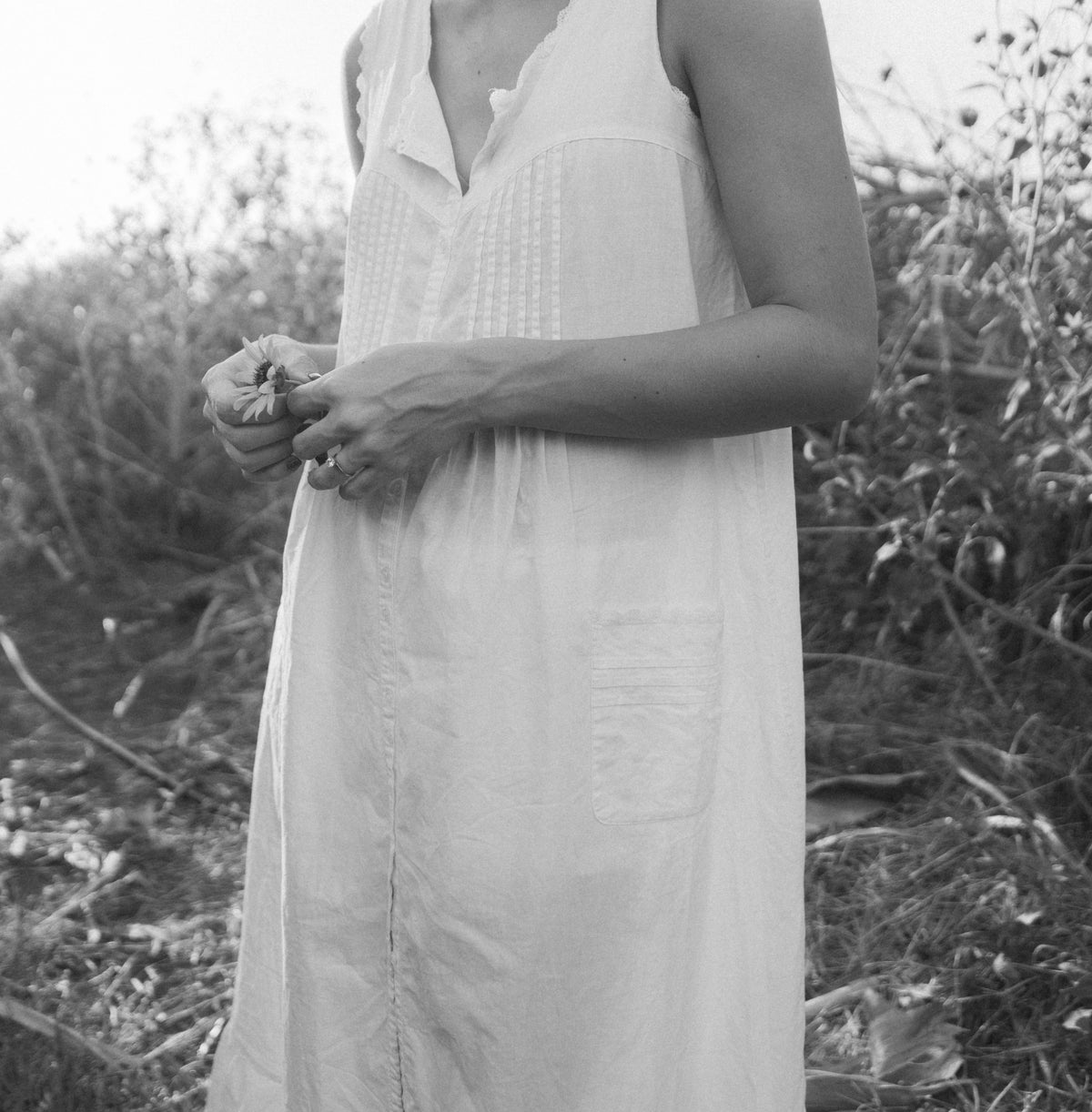 Woman in a long white dress holding a flower while standing among garden plants, photographed in black and white.