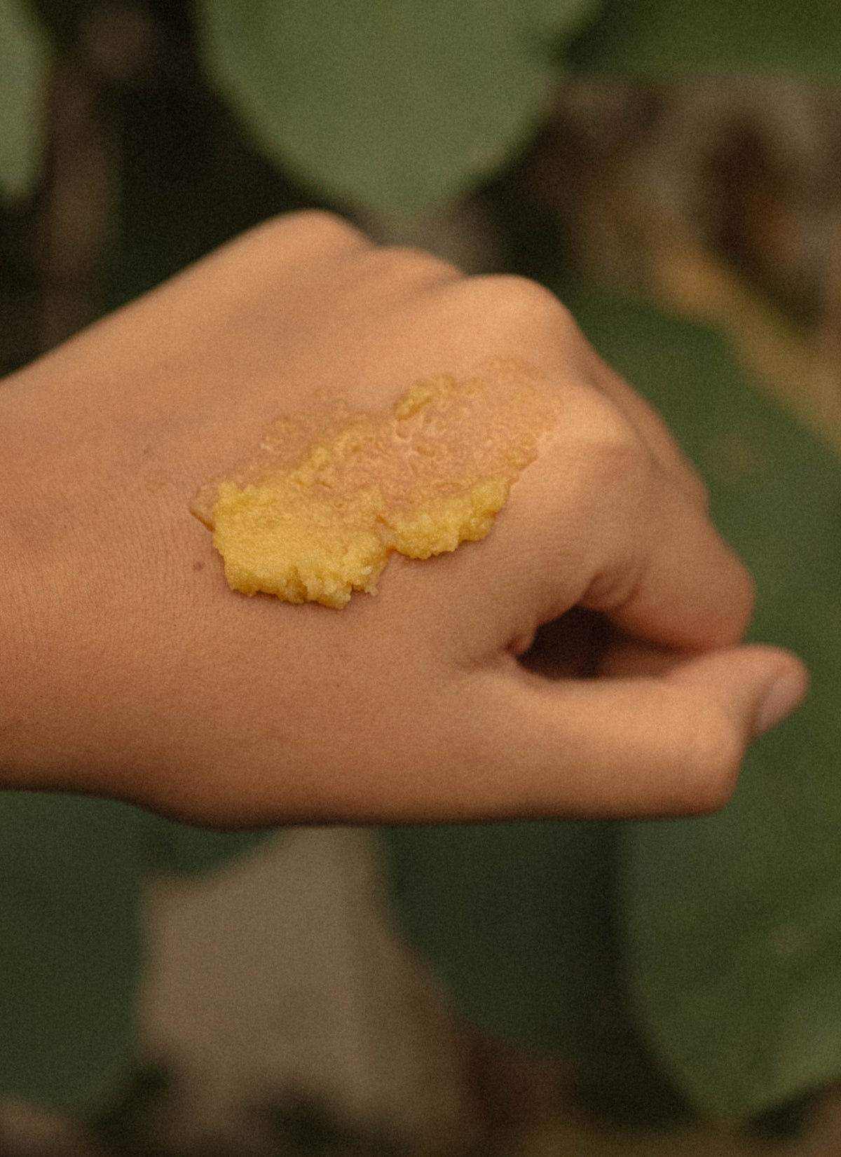 A close-up of a hand with a dollop of golden Salt Folk Rory Balm applied to the back of the hand, with a blurred green leafy background.