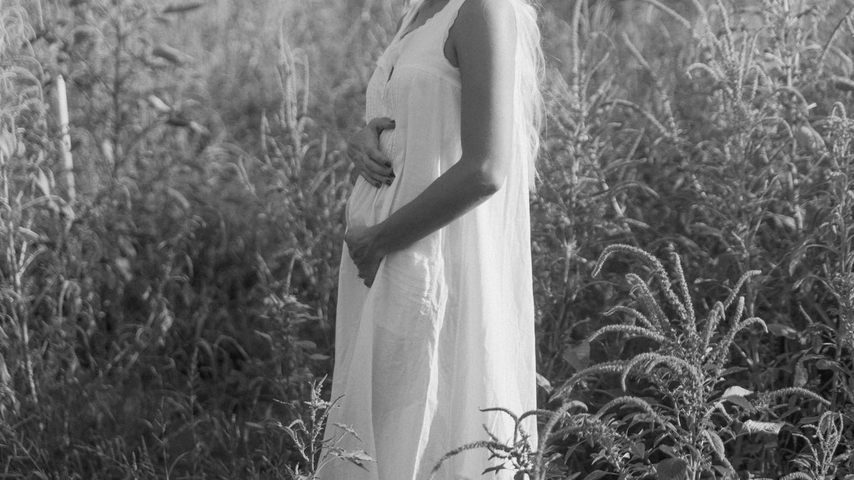 Pregnant woman in a long white dress gently resting her hands on her belly while standing among tall plants, photographed in black and white.