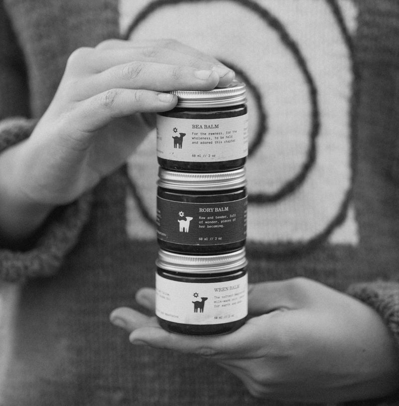 Hands holding a stack of three Salt Folk skincare balm jars against a knit sweater with a spiral design.