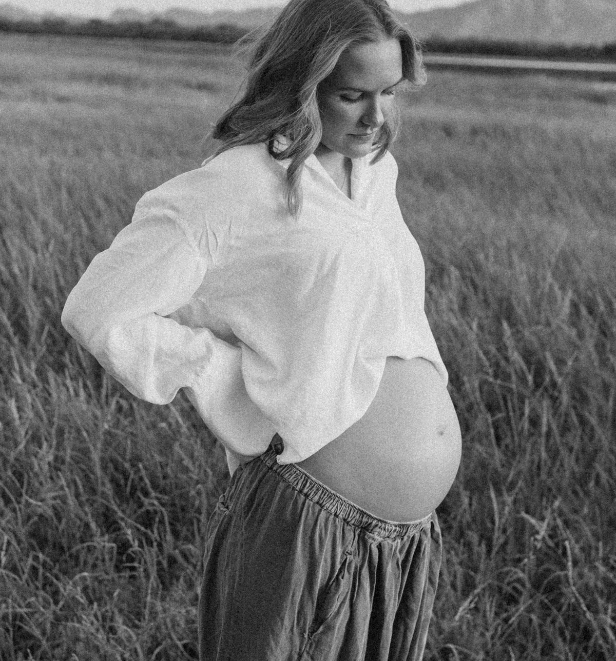 A pregnant woman standing in a grassy field, wearing a loose white long-sleeve top and a dark gray skirt.