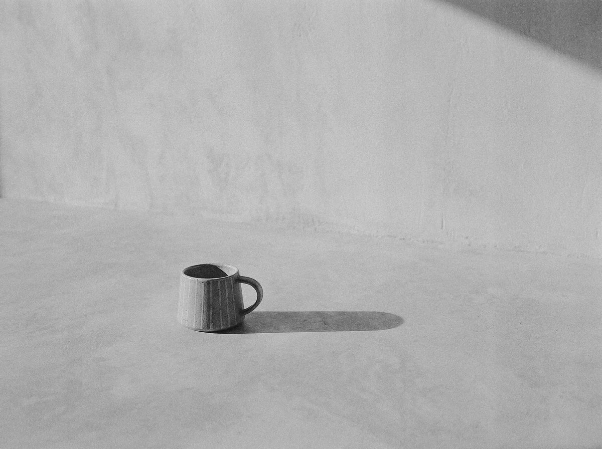 A single ceramic mug with a handle sits on a concrete floor against a textured plaster wall, casting a long shadow.