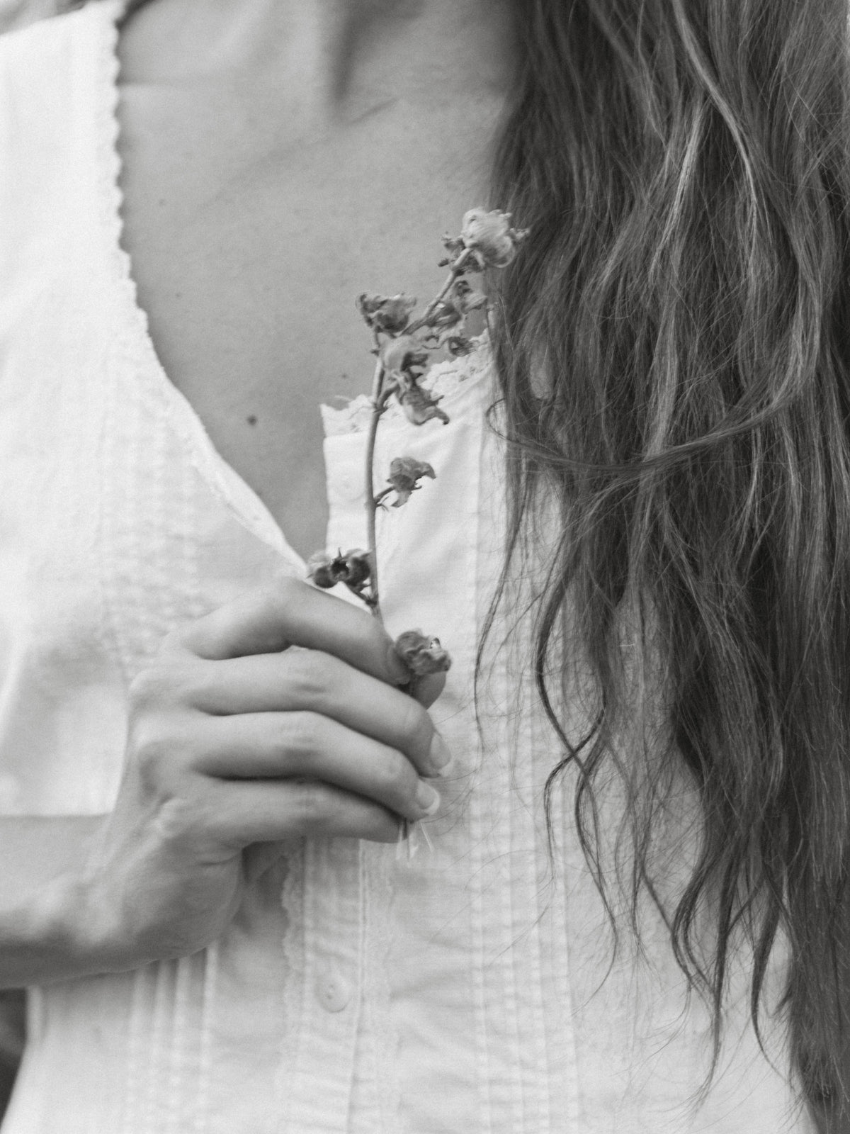 A close-up black and white photograph of a woman holding a small dried flower stem against her white textured blouse.